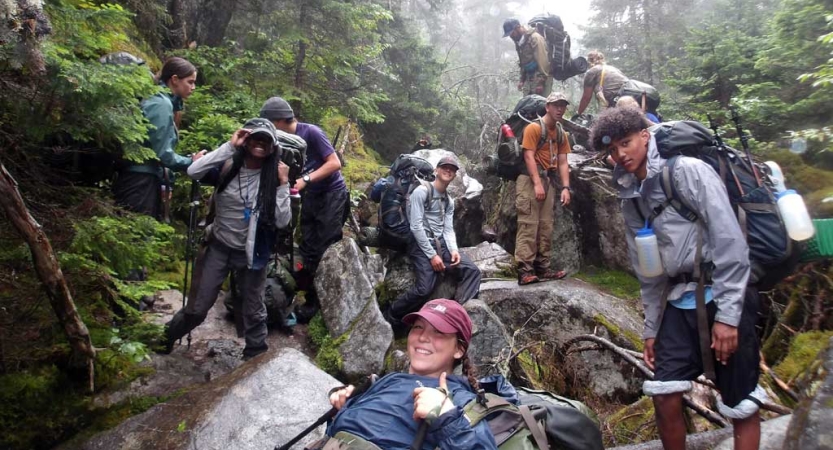 A group of students take a break from hiking and rest on rocks in a wooded area. 